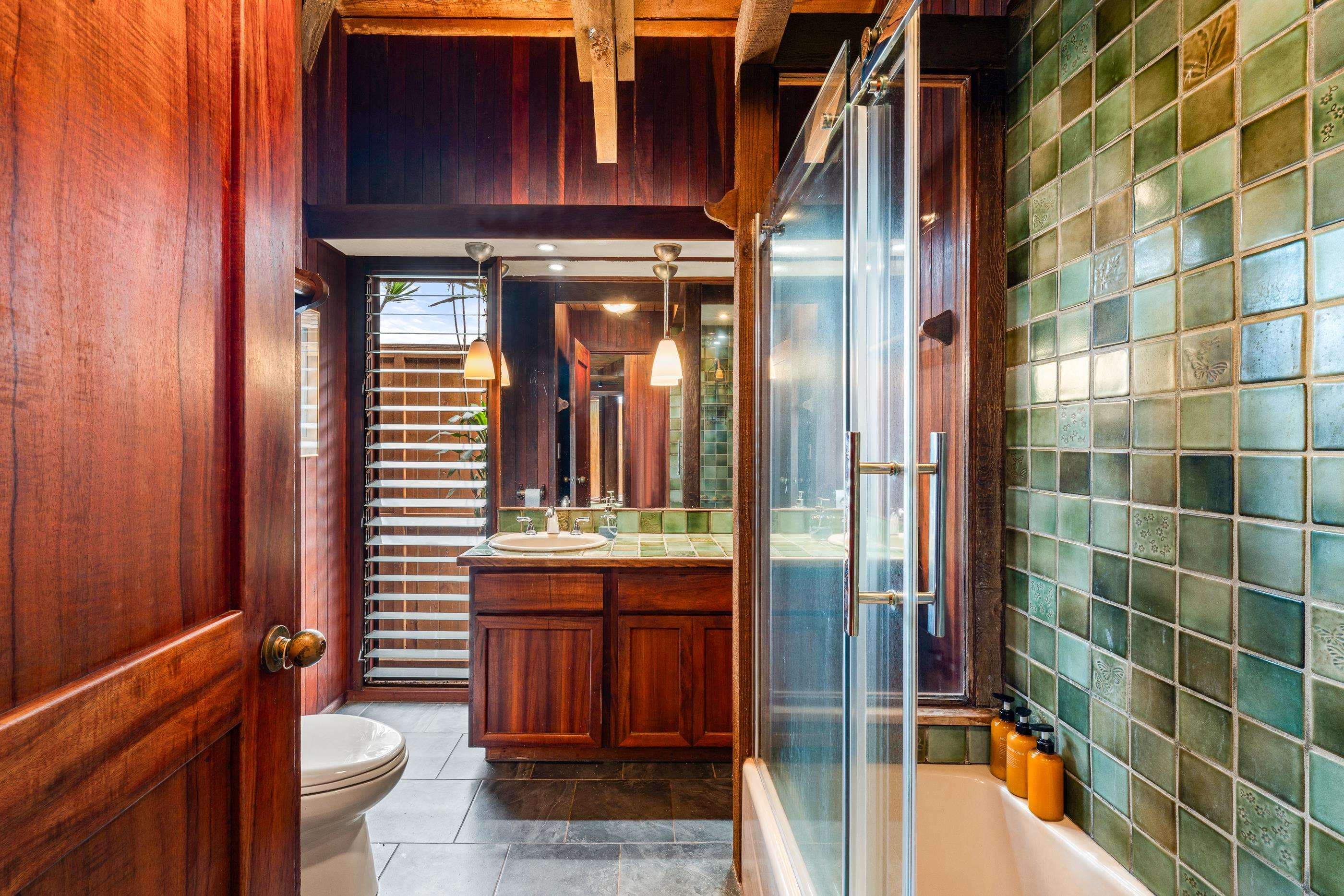 Bathroom in sea-glass tile, walk-in shower beside the wood vanity and louvered window
